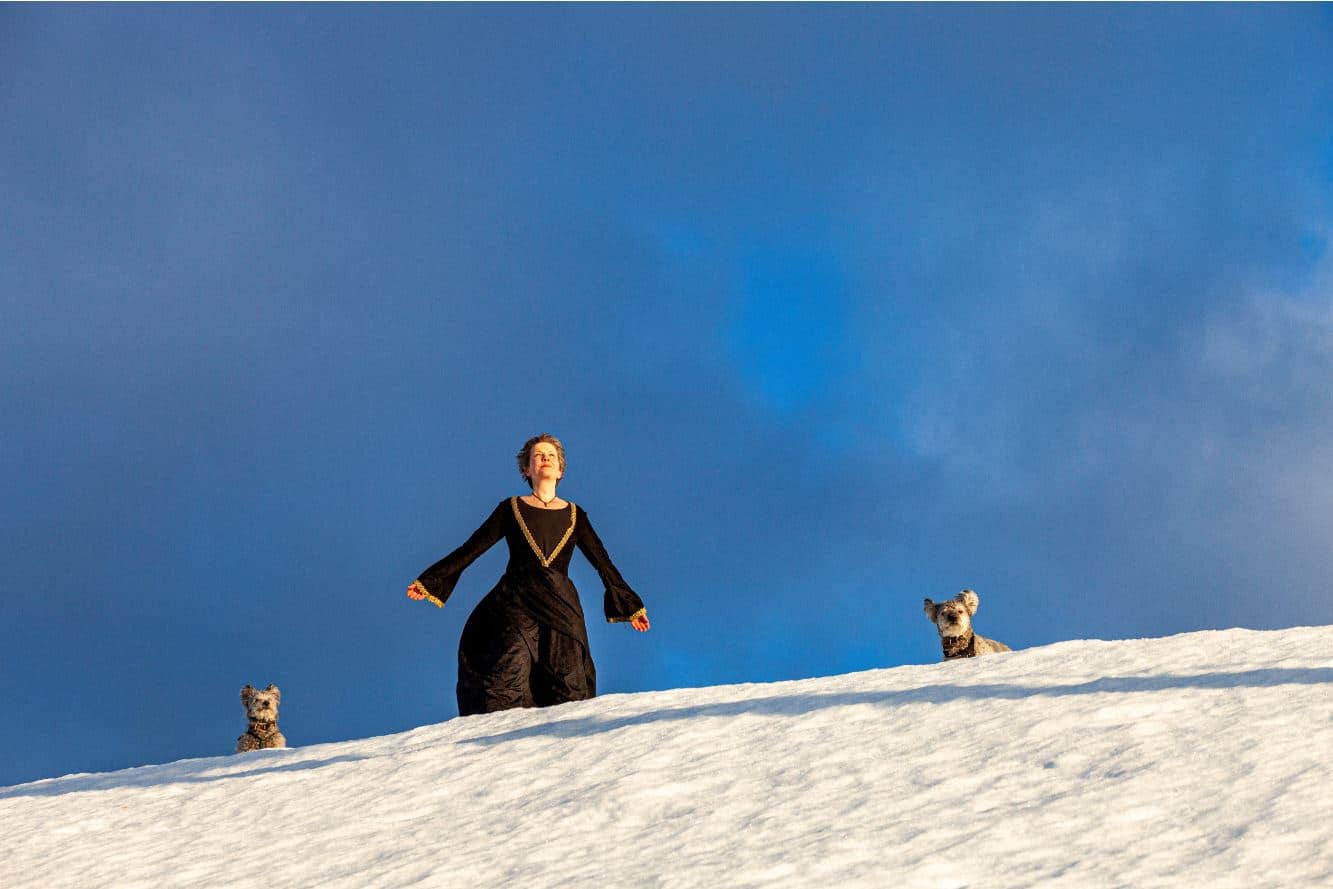 Sari Nyman seisoo hiekkadyynillä kädet sivuille levitettynä, vierellään kaksi koiraa.